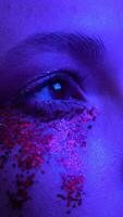 Close-up view of female face with shiny stage makeup with sparkles. Beauty night shot of woman eye illuminated blue purple color neon lighting. Selective focus. Part of series. Vertical handheld video