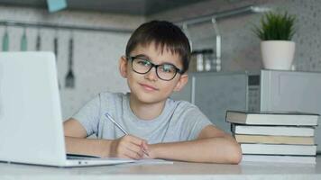 Attractive child with glasses does homework while at home sitting in the kitchen at the table using a laptop, textbooks, notebook. Portrait video