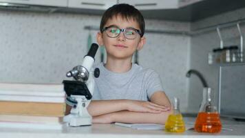 een schooljongen chemicus looks Bij de camera en glimlacht terwijl zittend Bij huis in de keuken De volgende naar een microscoop en leerboeken video