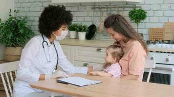 Afro american woman doctor in mask supporting a sick little girl sitting at the table at home. Family Doctor, Patient Support, Help at Home, Caring for the Sick. video