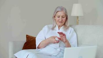 Skin Care, Senior Woman, Home Cosmetics, Elderly Age, Cosmetic Procedures. An elderly woman in a bathrobe during an online meeting uses a laptop and does beauty treatments video