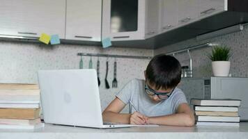 attraente bambino nel bicchieri insegna Lezioni mentre a casa seduta nel il cucina a il tavolo utilizzando un' computer portatile, libri di testo, taccuino. video