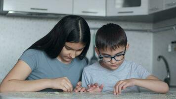 Cheerful Beautiful brother and sister are assembling a puzzle while sitting at a table at home in the kitchen. Close-up video