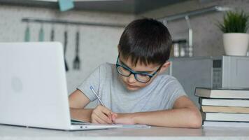 Attractive child in glasses teaches lessons while at home sitting in the kitchen at the table using a laptop, textbooks, notebook. Close-up video