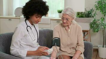 African american woman doctor checking blood pressure of senior woman while sitting on couch at home. Family Doctor, Patient Support, Help at Home, Caring for the Sick. video