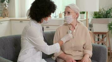 Afro american woman doctor in mask listens to the breathing of an elderly woman at home and supports her with her hand on her shoulder. Family Doctor, Patient Support, Help at Home video