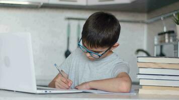 das Kind im Brille tut Hausaufgaben mit ein Laptop während Sitzung beim Zuhause im das Küche beim das Tabelle und schreibt mit ein Brunnen Stift im ein Notizbuch. online Technologie video