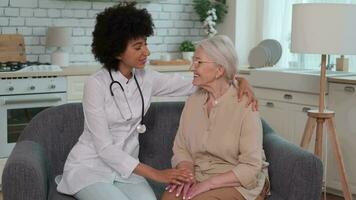 afro américain femme médecin les soutiens Sénior femme avec main sur épaule tandis que séance sur canapé. famille médecin, patient soutien, Aidez-moi à maison, soins pour le malade. video