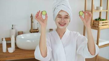 peau se soucier, après bain, produits de beauté à maison, caucasien femme, anti vieillissement. marrant caucasien femme dans une pansement robe et une serviette sur sa tête s'applique concombres à sa yeux pendant cosmétique procédures video