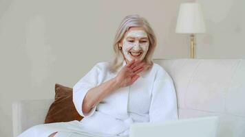 Haut Pflege, Senior Frau, Zuhause Kosmetika, Alten Alter, kosmetisch Verfahren. ein Alten Frau im ein kosmetisch Gesicht Maske kommuniziert mit online Technologien und ein Laptop während Sitzung auf das Couch video