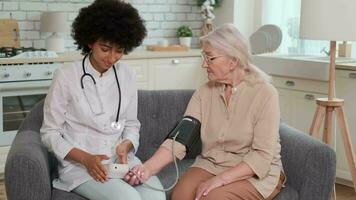 africano americano donna medico controllo sangue pressione di anziano donna mentre seduta su divano a casa. famiglia medico, paziente sostegno, Aiuto a casa, cura per il malato. video