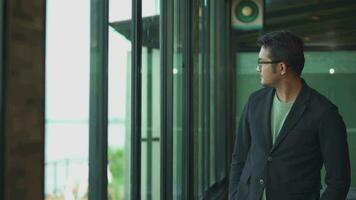 A handsome asia young stands window side by the window, he is dressed in a black suit, an office worker. video