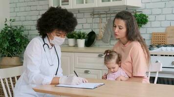 africano americano mujer médico en máscara escritura un prescripción mientras examinando un enfermo pequeño niña a hogar. familia doctor, paciente apoyo, ayuda a hogar, cuidando para el enfermo. video