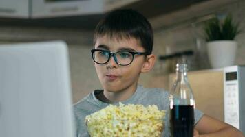 Stay at home, leisure at home, technologies for children, self isolation. Attractive boy in glasses looks at cartoons while sitting at home using a laptop and eating popcorn with soda. Portrait video
