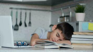 cansado colegial con lentes cayó dormido haciendo deberes sentado a un mesa siguiente a un ordenador portátil y libros de texto video