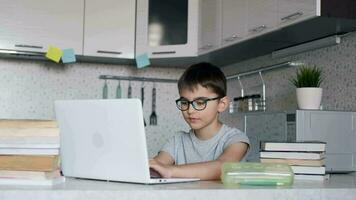 aantrekkelijk kind een schooljongen in bril geeft les lessen terwijl Bij huis zittend in de keuken Bij de tafel gebruik makend van leerboeken en een laptop. portret van een schooljongen. video