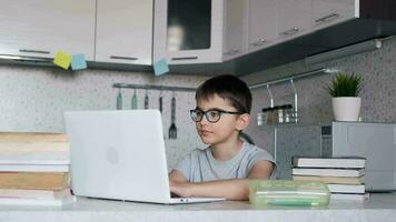 attraente bambino un' scolaro impara Lezioni mentre a casa seduta nel il cucina a il tavolo utilizzando libri di testo e un' il computer portatile. ritratto di un' scolaro. video