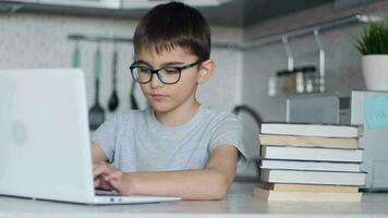 retrato. un niño en lentes haciendo deberes utilizando un ordenador portátil mientras sentado a hogar en el cocina a el mesa y mirando a el cámara. en línea tecnología video