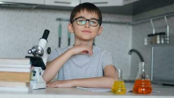 A schoolboy chemist looks at the camera and smiles while sitting at home in the kitchen next to a microscope and textbooks. Portrait video