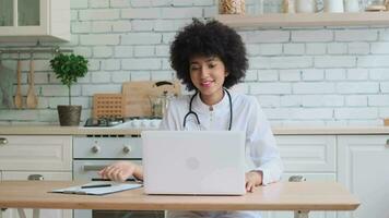 afro Amerikaans vrouw dokter in gewaad toepassingen laptop gedurende online overleg Bij huis terwijl zittend Bij tafel. familie dokter, geduldig steun, helpen Bij huis, zorgzaam voor de ziek. video