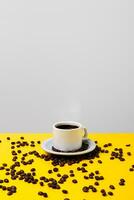 Coffee cup over bright yellow table and grey background and many beans spilled around it. photo