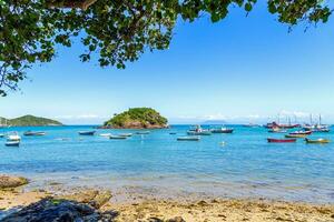 Seascape of Armacao Beach in Buzios, Rio de Janeiro, Brazil. Panoramic view of the bay. Boats and sailboats anchored in the sea. Tree leafs, clear water, blue sky, some rocks and a island. photo