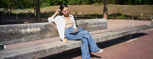 Smiling korean girl sitting in park with laptop, drinking takeaway coffee, enjoying sunny day, working or studying outdoors photo