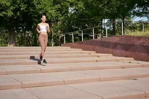 Portrait of young sportswoman running on street. Fitness girl jogging in park, workout outdoors, listening music in wireless earphones photo