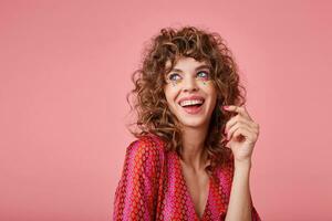 Beautiful young woman with curls laughing, being in a nice mood, standing over the pink background, wearing striped clothes with pattern, twisting a hair on a finger, looking away photo