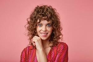 Flirty young woman looking at camera, biting a lip, looking coquettish, wearing striped pink and orange clothes with pattern, touching her hair, standing over the pink background photo