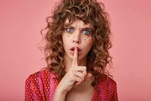 Pretty young woman with gloomy expression, frowning and keeping fore finger on lips, urges to calm down, not to produce noise, isolated over pink background photo