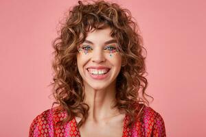 Portrait of beautiful young woman with curls, smiling cheerfully, showing her white teeth to camera, looking happy, standing over pink background photo