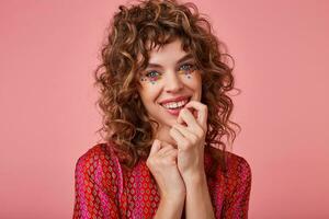 Indoor shot of young lovely woman with curls, smiling widely and touching her face, looking pleased and happy, isolated over pink background photo