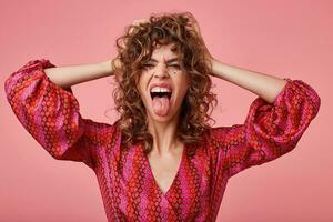 Portrait of excited young woman, screaming loud and sticking out tongue, covering ears with hands, standing over pink background. wearing striped pink and orange clothes with pattern photo