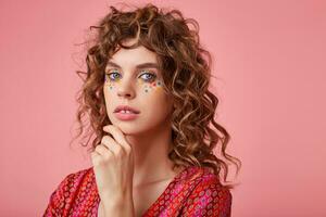 Young pretty woman over the pink background keeping hand under chin, wearing striped pink and orange clothes with pattern, looking calm to the camera with light smile photo