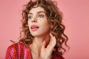 Indoor shot of attractive young woman with curls, looking away and touching her neck, has dreaming and romantic look, standing over the pink background photo