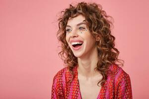 Pretty young curly lady looking away, being in nice mood, laughing, standing over pink background, wearing striped pink and orange clothes with pattern. Positive emotions concept photo