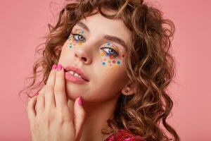 Close-up shot of attractive coquettish young woman with romantic curls, looking flirty at camera, touching her lips, standing over the pink background photo