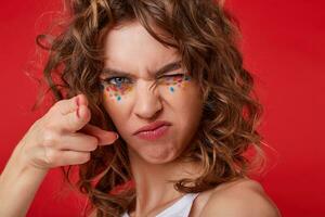 Portrait of young curly woman with closed eye, stands over red background, makes gun from her hand, looks serious, squints and threatens with a gun photo