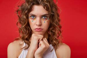 Pretty young woman with light-brown curly hair standing over the red background, looking offended and confused, clenching fists near to face photo