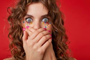 Shocked curly woman stands in front of camera with eyes wide open, crosses palms on her mouth, looks scared, shooted on red background photo