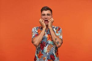 Indoor shot of young bewildered man with short haircut, wearing glasses and flowered shirt, making grimace and keeping hands on cheeks, posing on orange background photo
