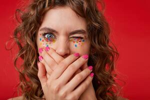 retrato de joven Rizado mujer con cruzado palmas en su labios, mirando a el cámara con uno cerrado ojo, en pie terminado el rojo fondo, muestra asco, siendo confuso foto