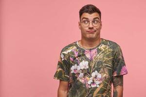 Young attractive man standing in front camera over the pink background wih raised eyebrows, looking away, wearing glasses and printed t-shirt, seems bored and indifferent photo