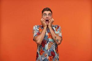 Portrait of young guy standing with wide opened eyes and mouth, screaming in fear, wearing glasses and flowered shirt, standing over orange background photo