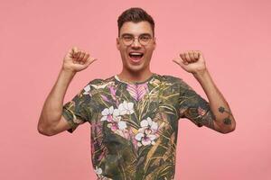 Photo of handsome guy with short cut and opened mouth, pointing thumbs, looking pleased with himself, wearing glasses and printed t-shirt, standing over pink background