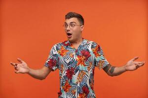 Surprised young man in floral shirt shrugs with opened mouth, standing over orange background, wearing glasses, looking away photo