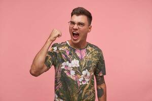 Portrait of handsome guy with glasses, raising a fist and shouting loudely, looking flurried and excited, posing on pink background photo