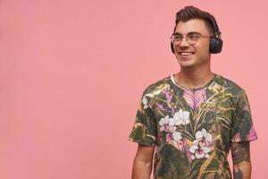 sonriendo de pelo corto hansome chico mirando lejos, siendo positivo y en bonito ánimo, disfrutando el música en auriculares, posando terminado rosado antecedentes foto