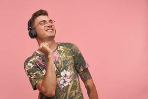 Indoor shot of satisfied young guy in flowered t-shirt with headphones, taking pleasure in musical track, standing over pink background photo